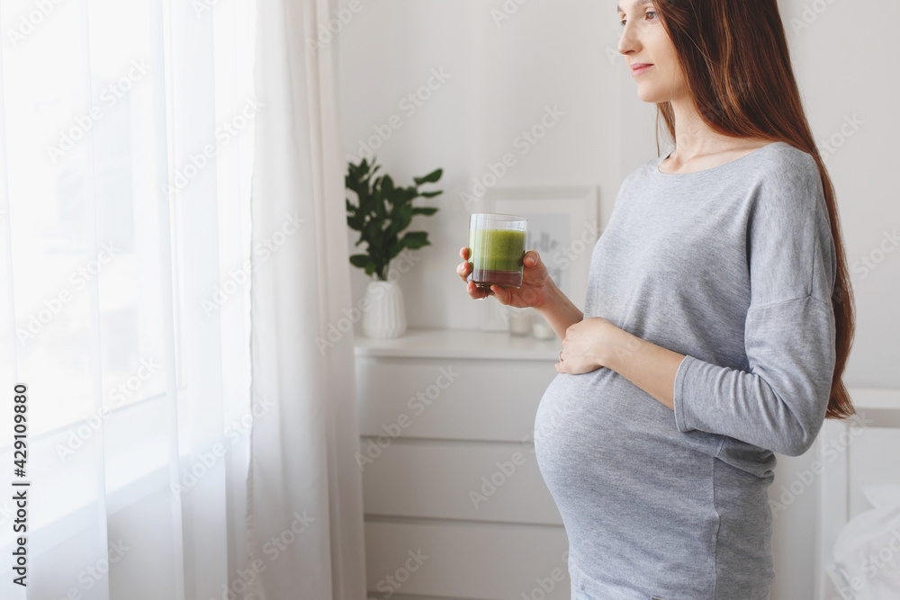 Young beautiful pregnant woman standing at home near the window and drinking a green smoothie. Young beautiful pregnant woman standing at home near the window and drinking a green smoothie.