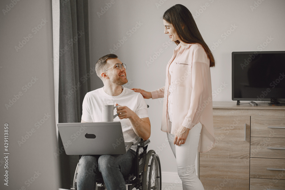 Young man in wheelchair using laptop and girlfriend hugging him at home Young man in wheelchair using laptop and girlfriend hugging him at home