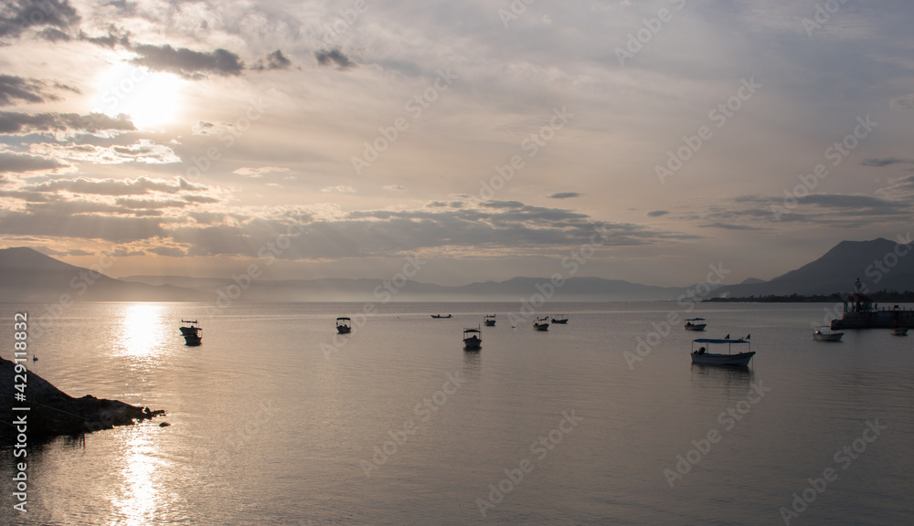 Boats sitting on the silvery surface of Lake Chapala at dusk with rays of sun through the clouds from the Malecon de Chapala, Mexico