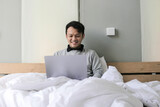 Happy Asian man is working with his laptop on his bed. Concept of freelancer successful lifestyle.