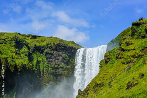 Tourists overlooking Iceland's Skogafoss waterfall surrounded by blue sky and greenery