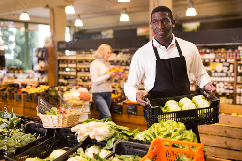 African male grocery worker in apron arranging fresh vegetables on shop counter