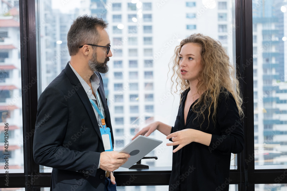 Business woman looking at man and discuss together standing beside windows in the modern high level office building. Man holding tablet. Teamwork, brainstorm and business concept. Business woman looking at man and discuss together standing beside windows in the modern high level office building. Man holding tablet. Teamwork, brainstorm and business concept.