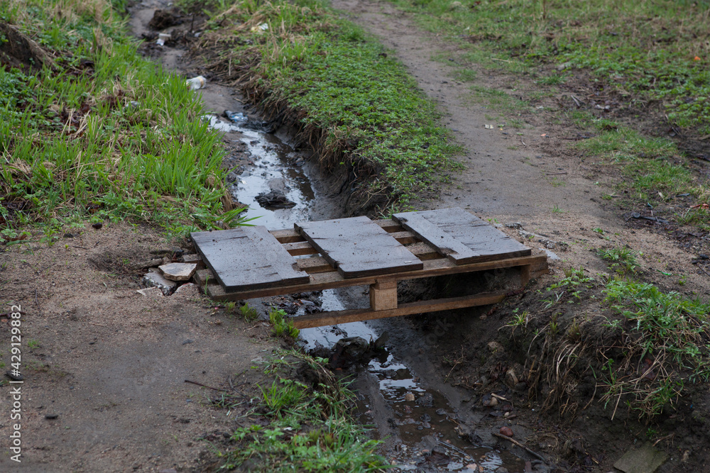 Fototapeta premium A small homemade wooden bridge over a muddy, smelly puddle or stream in a slum. Unkempt area, outskirts of the city.