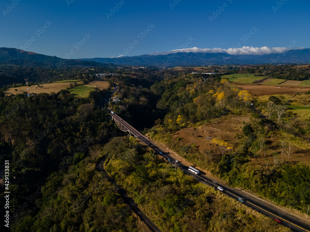 Beautiful aerial view of a bridge inside the yellow trees of the forest of Costa Rica, with the impressive mountains in the back 