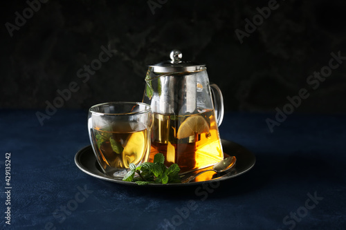 Teapot and glass of tasty tea with lemon and mint on dark background