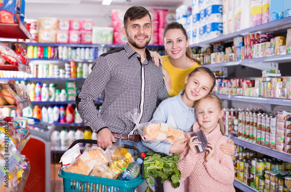 Cheerful family with two children holding full basket after shopping in food store