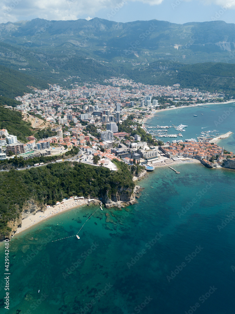 Aerial view of Budva, Montenegro. Mogren beach, mountainous landscape, turquoise emerald transparent water, stones on seabed. Old town with orange roofs, port with boats. Balkans travel destination