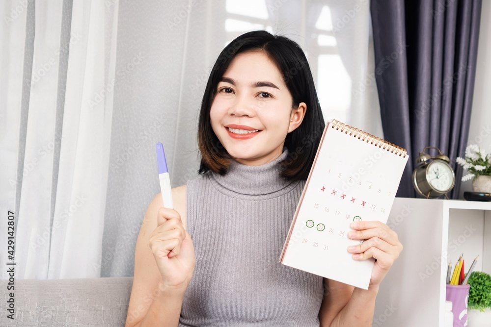 smiling Asian woman hand holding pregnancy test and calendar showing mark on date menstrual cycle planning for ovulation day smiling Asian woman hand holding pregnancy test and calendar showing mark on date menstrual cycle planning for ovulation day