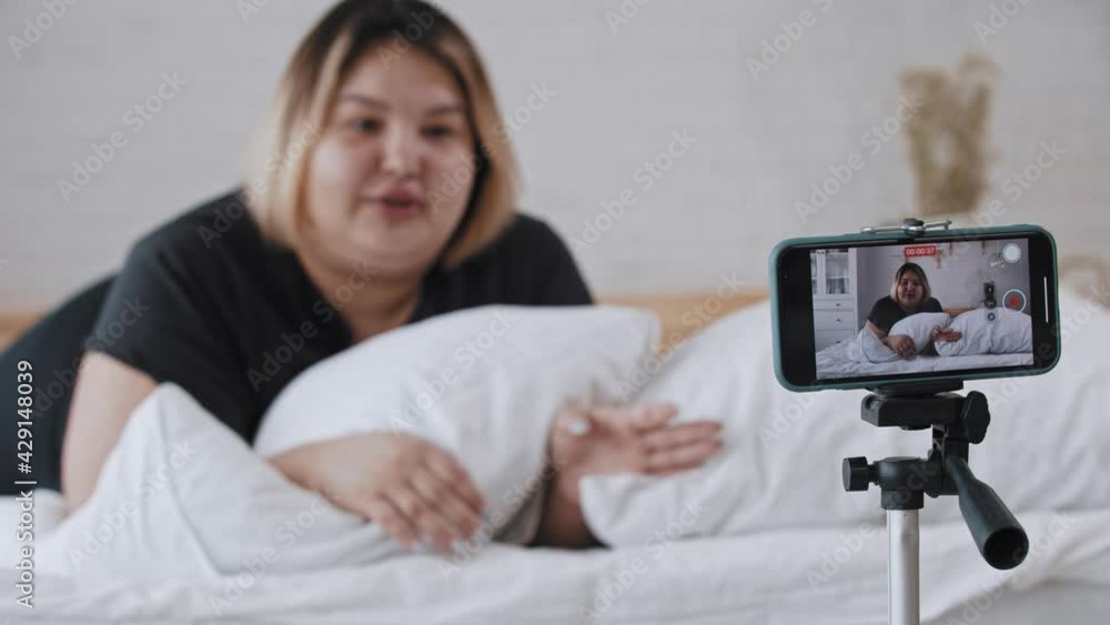 A confident overweight woman feminist lying in bed and talking on a video for her followers about body positivity A confident overweight woman feminist lying in bed and talking on a video for her followers about body positivity
