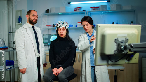 Team of medical researchers testing nervous system of woman patient, looking together at brain scan on computer monitor. Doctors examining evolution of treatment against diseases in equipped lab