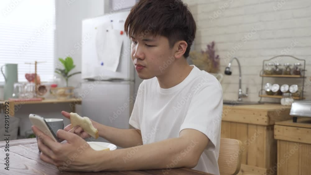 asian boy sitting alone with a smartphone is phubbing while drinking tea and eating toast in the morning at home. asian boy sitting alone with a smartphone is phubbing while drinking tea and eating toast in the morning at home.