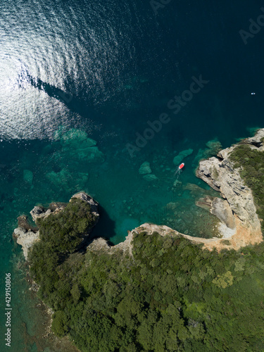 Fototapeta Naklejka Na Ścianę i Meble -  Aerial view of red boat sailing near steep stone cliff with green forest above transparent emerald water of Adriatic sea. Stones on seabed. Unspoiled nature of Montenegro coast. Budva scenic landscape