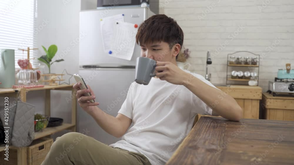 portrait japanese young guy is watching online videos on his smartphone while sitting at dining table and sipping coffee in leisure. portrait japanese young guy is watching online videos on his smartphone while sitting at dining table and sipping coffee in leisure.