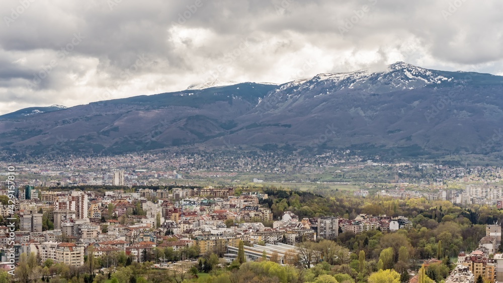 Aerial view of urban landscape with many administrative and residential buildings, green park and mountain background, cloudy sky. Sofia, the capital city of Bulgaria, East Europe. Panoramic sight.