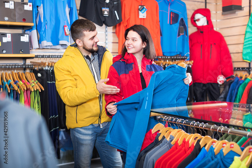 Young positive couple choosing track jacket in sports clothes shop