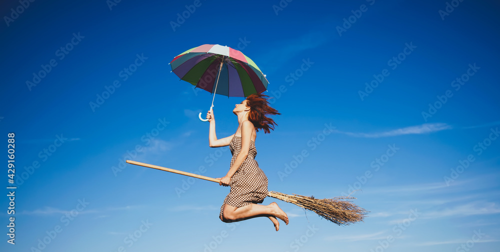 Young red-haired witch on broom flying in the sky with umbrella