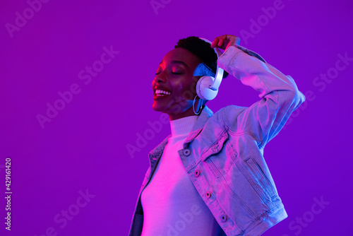 Young African American woman wearing headphones listening to music and dancing in futuristic purple cyberpunk neon light background photo