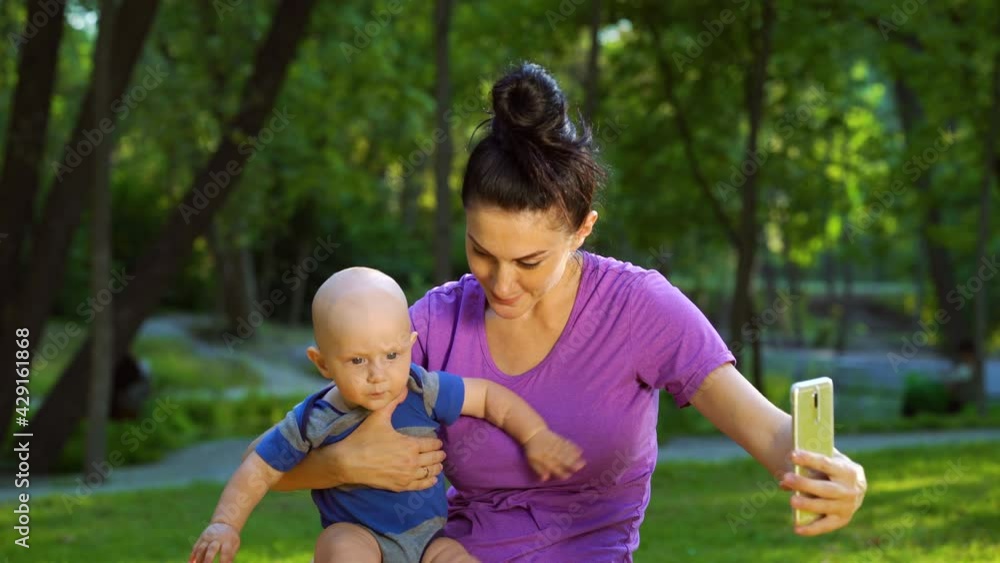 Woman with hair bun holding baby and recording video with smartphone in park, blurred people walking on background. Mother streaming for family blog. Concept of gadget Woman with hair bun holding baby and recording video with smartphone in park, blurred people walking on background. Mother streaming for family blog. Concept of gadget