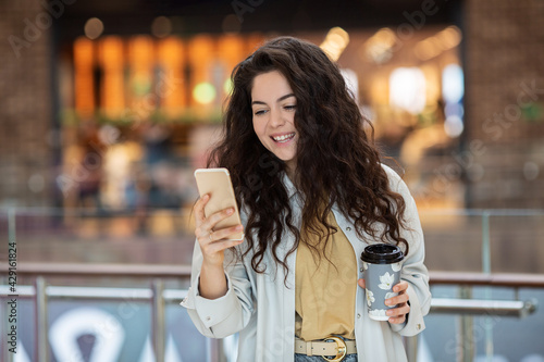 Young cheerfful woman using smartphone and drinking take away coffee in paper cup. Stylish woman at cafe indoors. photo