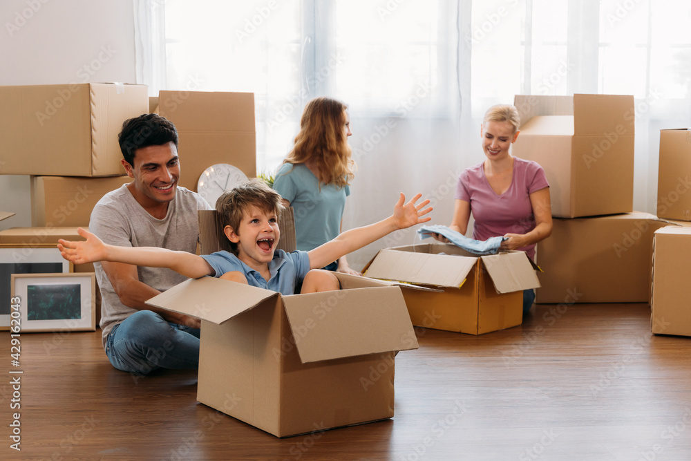 Playful little boy sitting in cardboard enjoying with young caucasian parents and sister unpacking stuff from carton boxes in new home Playful little boy sitting in cardboard enjoying with young caucasian parents and sister unpacking stuff from carton boxes in new home