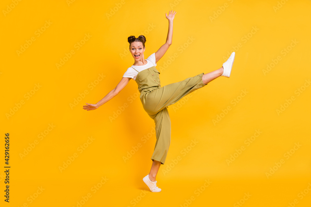 Photo of funny adorable lady dance open mouth raise leg wear green overall shoes isolated yellow background