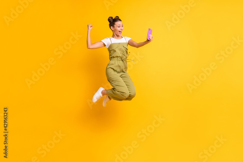 Photo of crazy lady jump hold telephone raise fist open mouth wear green overall shoes isolated yellow background