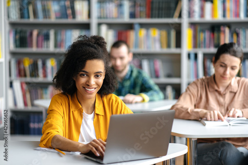 Students sit at their desks in the library during a lecture. African American curly pretty female student sits at a table with a laptop during a lesson, looks at the camera, friendly smiling