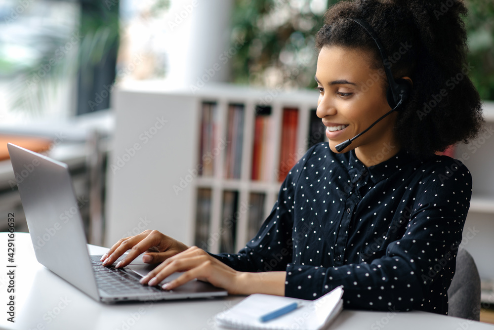 African American beautiful young woman wearing a headset, sitting at a work desk, using a laptop, working online, surfing the Internet, chatting with colleagues or clients, answers to email, smiling African American beautiful young woman wearing a headset, sitting at a work desk, using a laptop, working online, surfing the Internet, chatting with colleagues or clients, answers to email, smiling