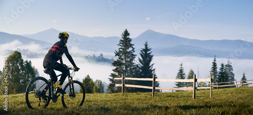 Fototapeta Naklejka Na Ścianę i Meble -  Man riding bicycle on grassy hill and looking at beautiful misty mountains. Male bicyclist enjoying panoramic view of majestic mountains during bicycle ride. Concept of sport, bicycling and nature.