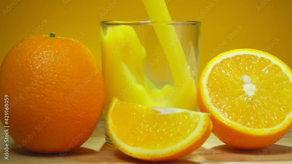 Fresh delicious orange juice is poured in a glass next to large ripe oranges cut into slices on the yellow background in slow motion Fresh delicious orange juice is poured in a glass next to large ripe oranges cut into slices on the yellow background in slow motion