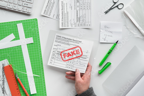 Fake covid vaccination record card. Covid scams. Forged health certificate with false vaccination record against covid-19. Blank card, pen, paper knife,laminator. Top view, flat lay on white table.