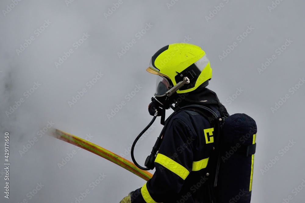 Naklejka premium A firefighter in a helmet and with a breathing apparatus is shrouded in smoke holding a hose during firefighting