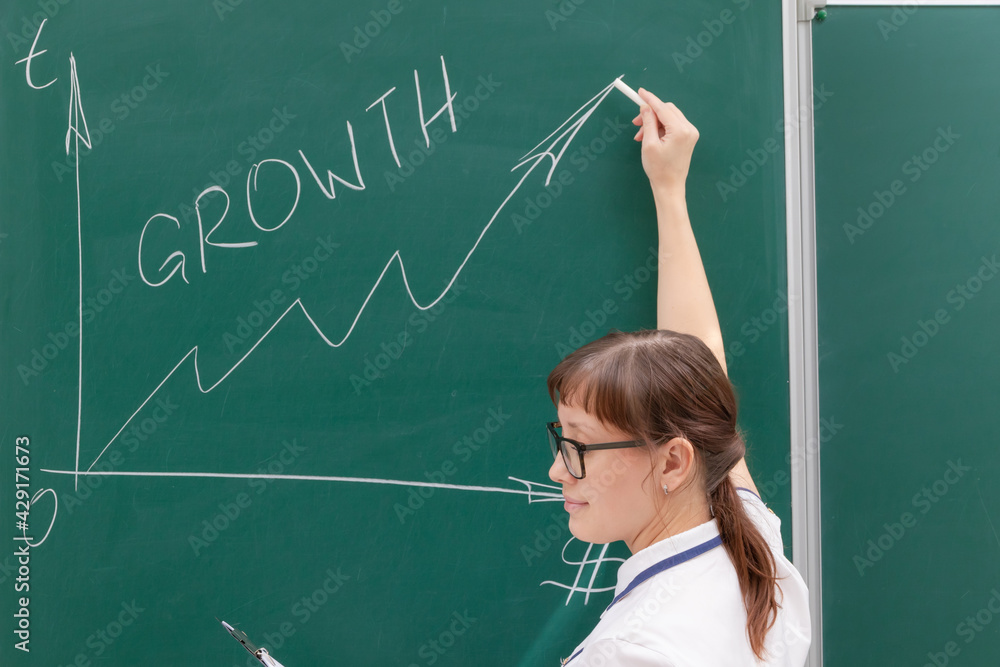 young woman business coach in a white blouse and glasses at the chalkboard in the office draws a profit graph. close-up