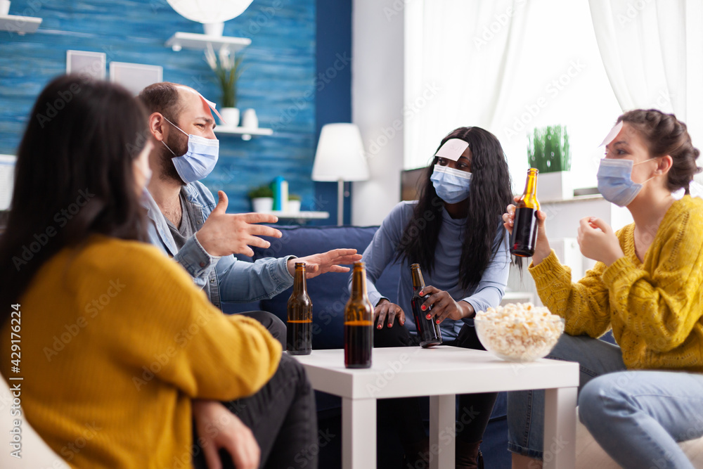 Funny friends with sticky notes on foreheads enjoying name game gesturing  during new normal party wearing face mask keeping social distancing  preventing covid19 spread. People enjoying bottles of beer Stock Photo |