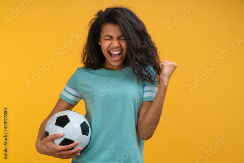 portrait of cute dark skinned soccer fan girl with a ball in hand celebrating favourite team victory making winner's gesture, clenching her fist and screaming yes, isolated on orange yellow background © wpadington