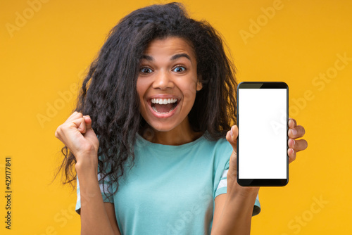 Beautiful smiling dark skinned girl showing mobile phone with blank screen directly to the camera and clenching her fist in winner's gesture, isolated on bright colored yellow background