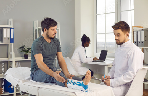 Doctor talking to male patient and prescribing treatment for his foot injury. Worried young man with ankle sprain consulting orthopedic surgeon sitting on examination bed at modern hospital or clinic