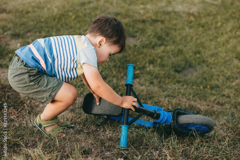 Fototapeta premium Caucasian small boy is getting up the bike after falling down in a field on grass