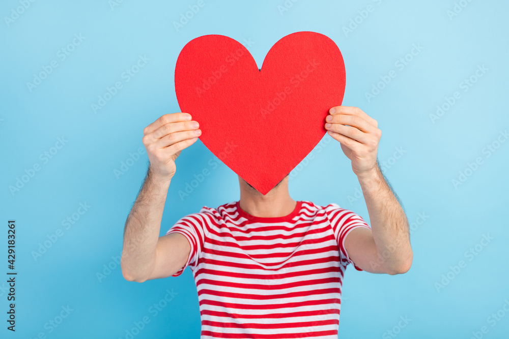 Photo of sweet shy young man dressed striped t-shirt red paper heart cover face head empty space isolated blue color background Photo of sweet shy young man dressed striped t-shirt red paper heart cover face head empty space isolated blue color background