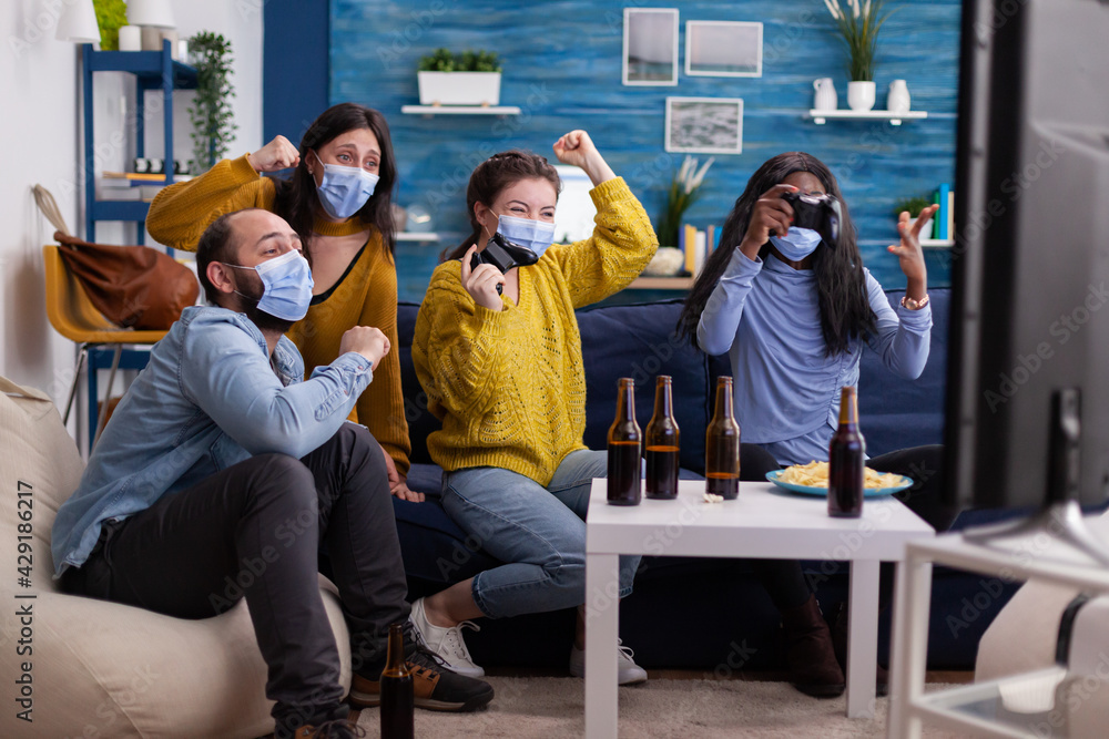Multiethnic people celebrate video game victory in home living room with joystick wearing face mask keeping social distancing in time of corona outbreak. Diverse friends enjoying beer and chips.