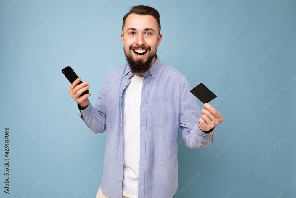 Photo of good looking attractive smiling brunet unshaven young man wearing casual blue shirt and white t-shirt isolated over blue background wall holding credit card and using mobile phone making