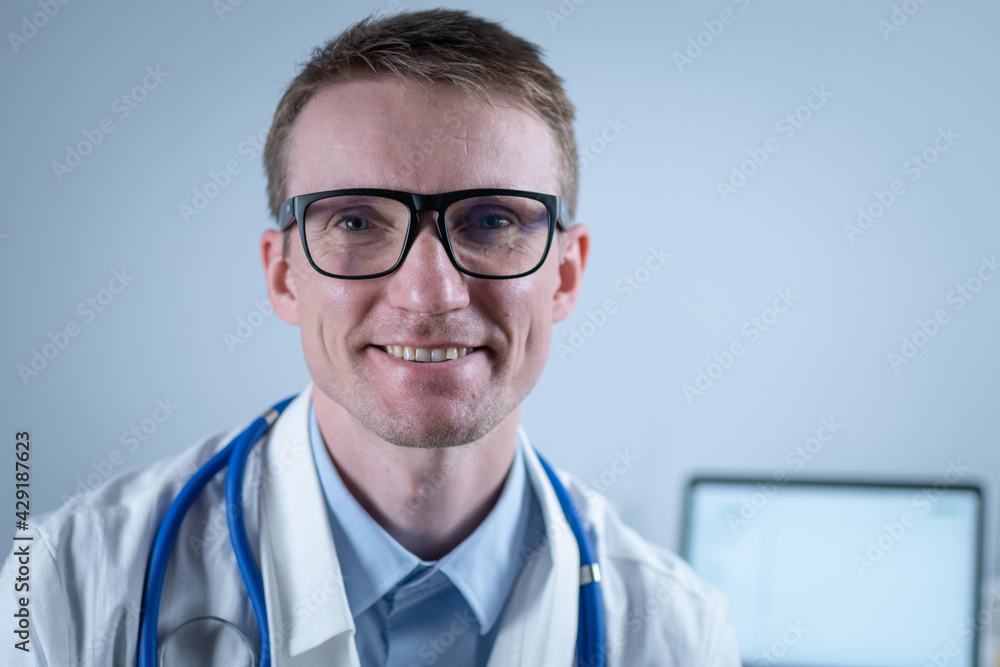 Portrait of adult handsome family doctor in medical office. Successful physician in white lab coat looking at camera and smiling in hospital clinic. Young general practitioner wearing glasses smiling Portrait of adult handsome family doctor in medical office. Successful physician in white lab coat looking at camera and smiling in hospital clinic. Young general practitioner wearing glasses smiling