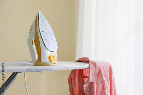 A single clean freshly laundered pink shirt and electric iron on pressboard at home. Clothing on ironing board, full length window with white curtains. Copy space, close up, top view, background