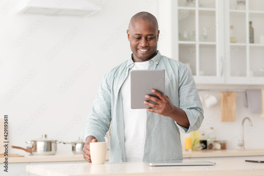 Positive african american mature man reading news on digital tablet and drinking coffee in kitchen