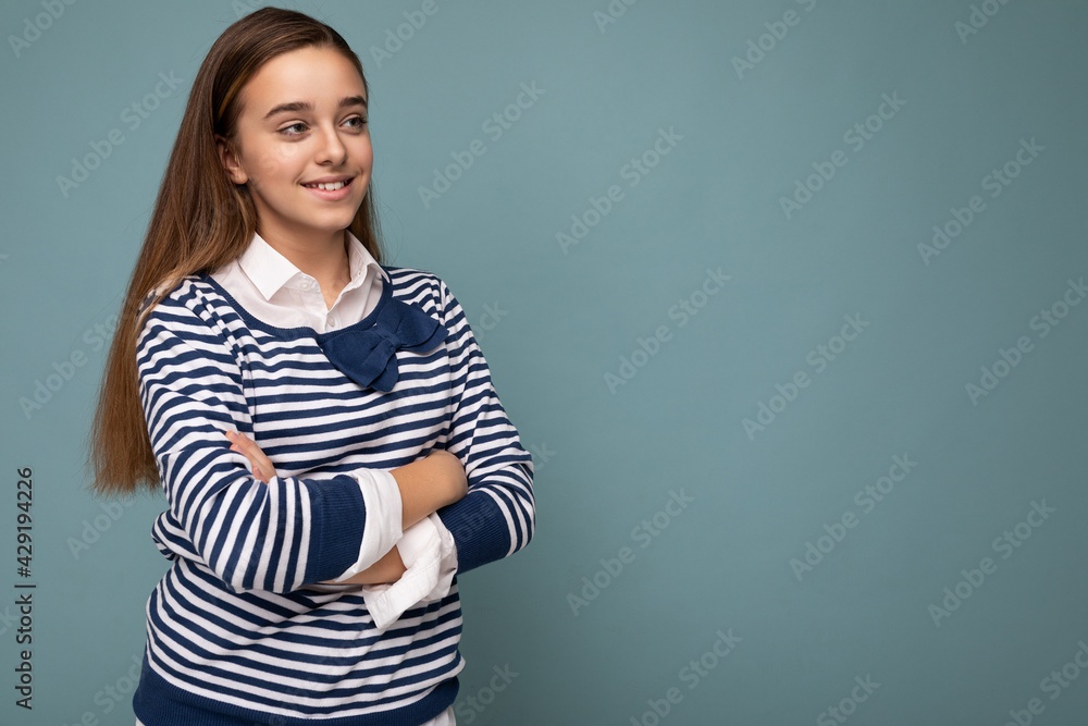 Shot of beautiful positive smiling brunette little girl wearing striped longsleeve standing isolated over blue background wall crossing hands and looking to the side