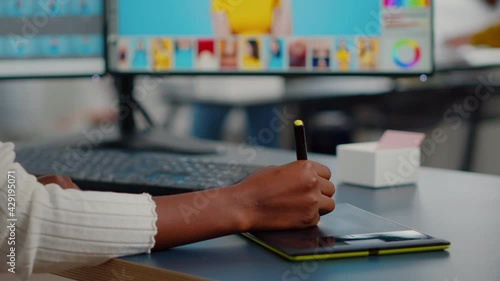 Close up of african photo retoucher holding stylus pen editing customer photo using computer with two displays. Graphic editor retouching assets drawing on graphic tablet in creative agency office