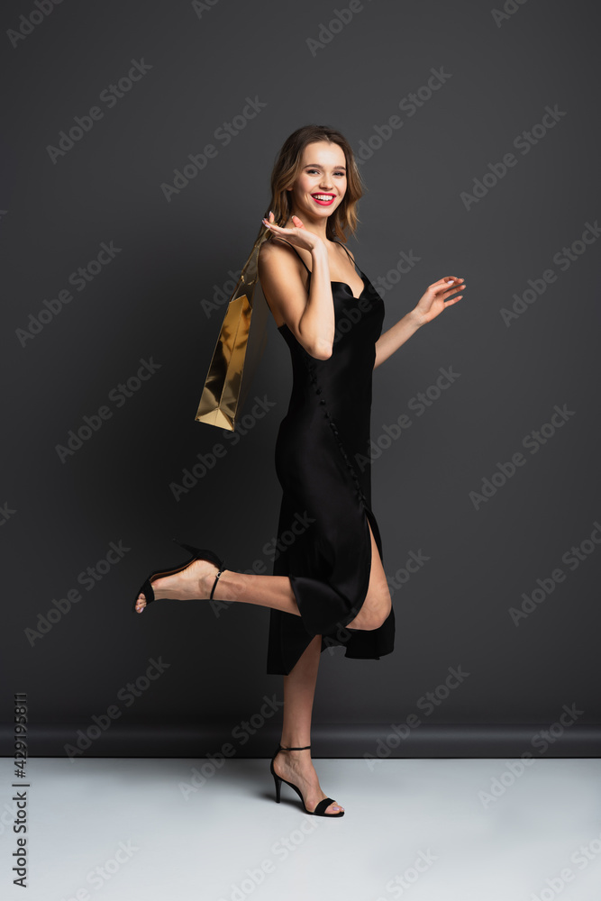 full length of cheerful young woman in black slip dress holding shiny shopping bag on grey
