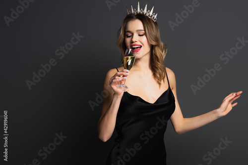 positive young woman in black slip dress and tiara holding glass of champagne on grey