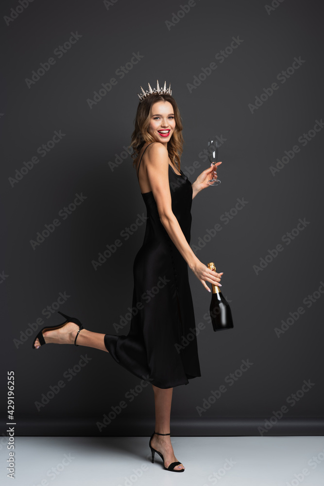 full length of happy woman in black slip dress and tiara with diamonds holding bottle of champagne and glass on grey